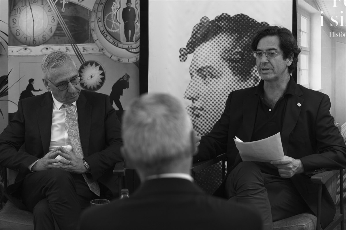 Dos participantes conversan en un acto público ante un retrato de gran formato.