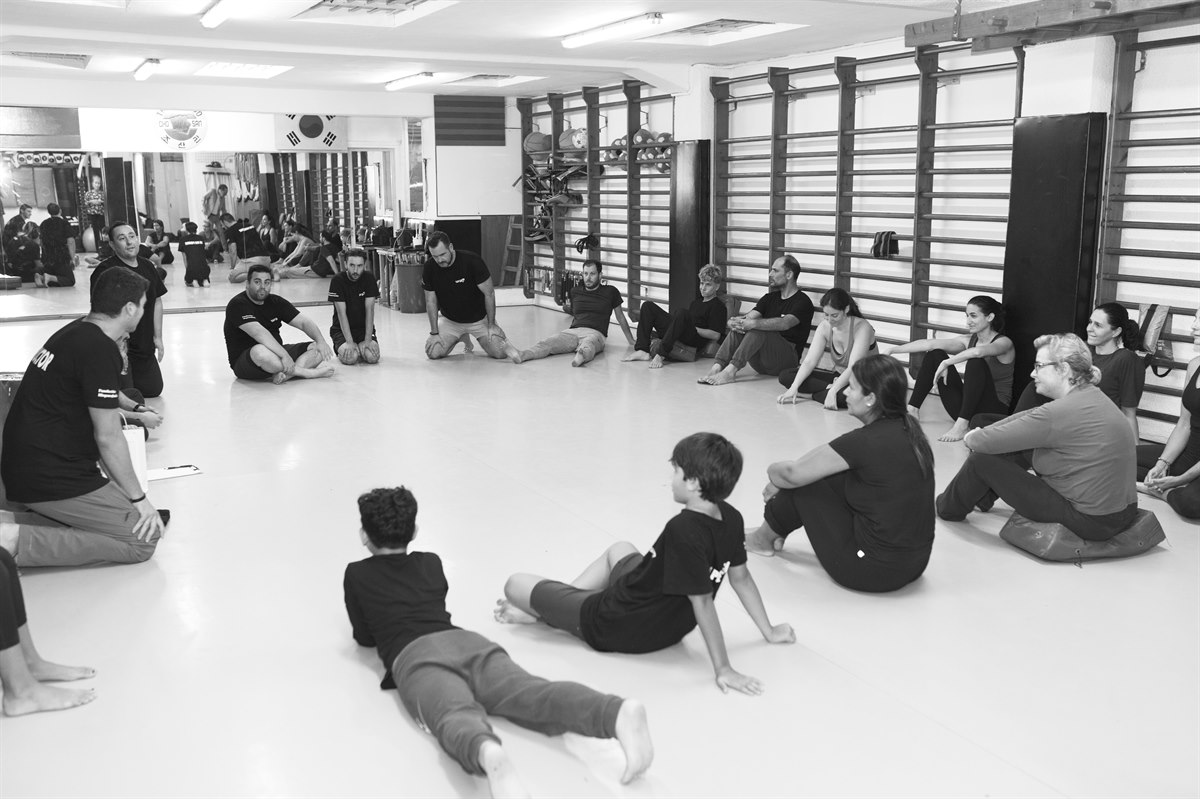 Grupo sentado en el suelo de una sala durante una sesión de defensa personal.