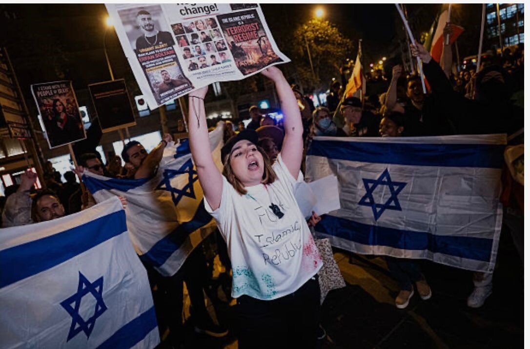 Manifestación nocturna: una participante alza un cartel que denuncia ejecuciones en Irán, rodeada de banderas de Israel y de Irán.