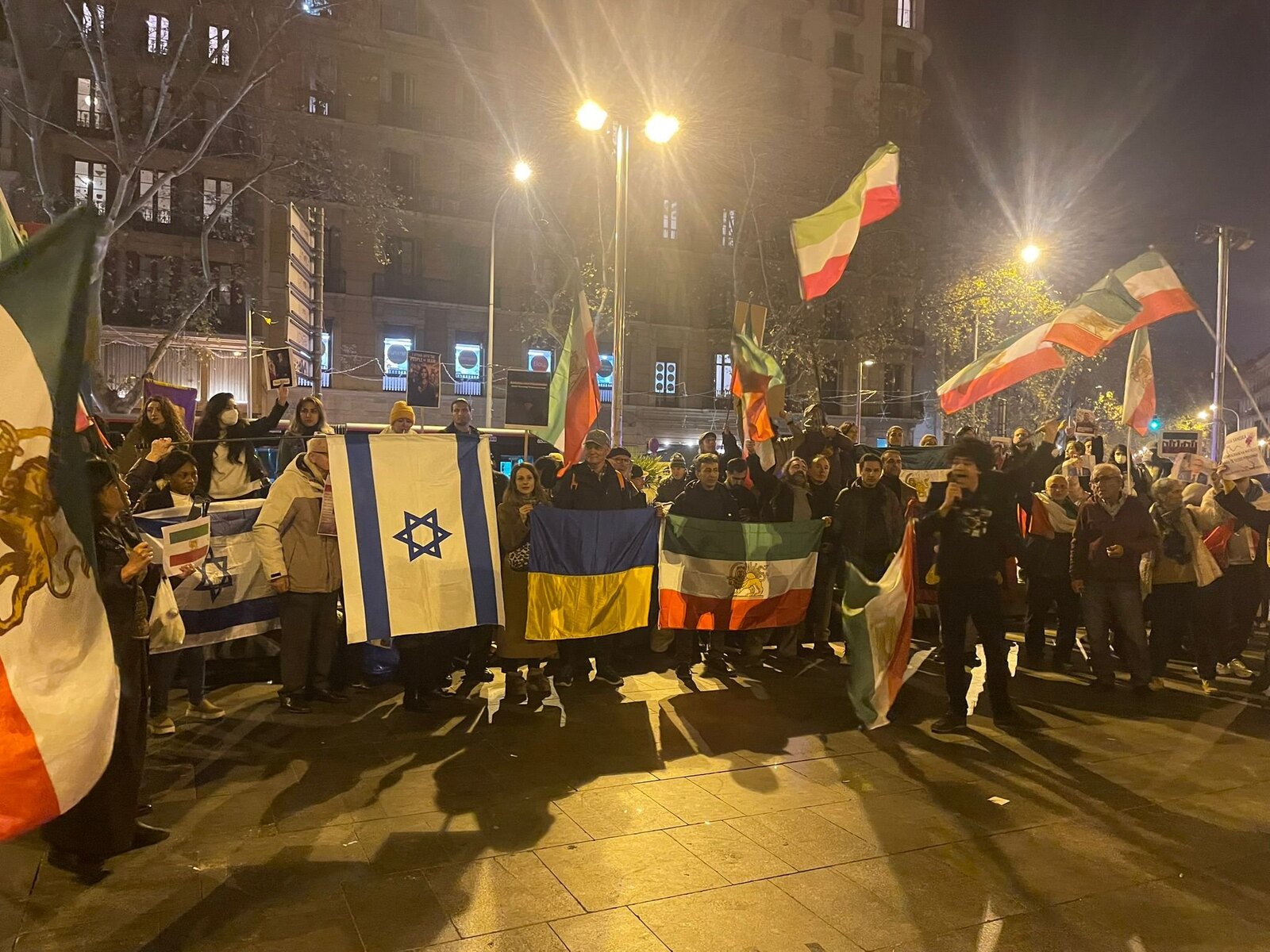 Concentración nocturna en una avenida de Barcelona: un grupo sostiene banderas de Irán, de Israel y de Ucrania frente a los edificios iluminados.