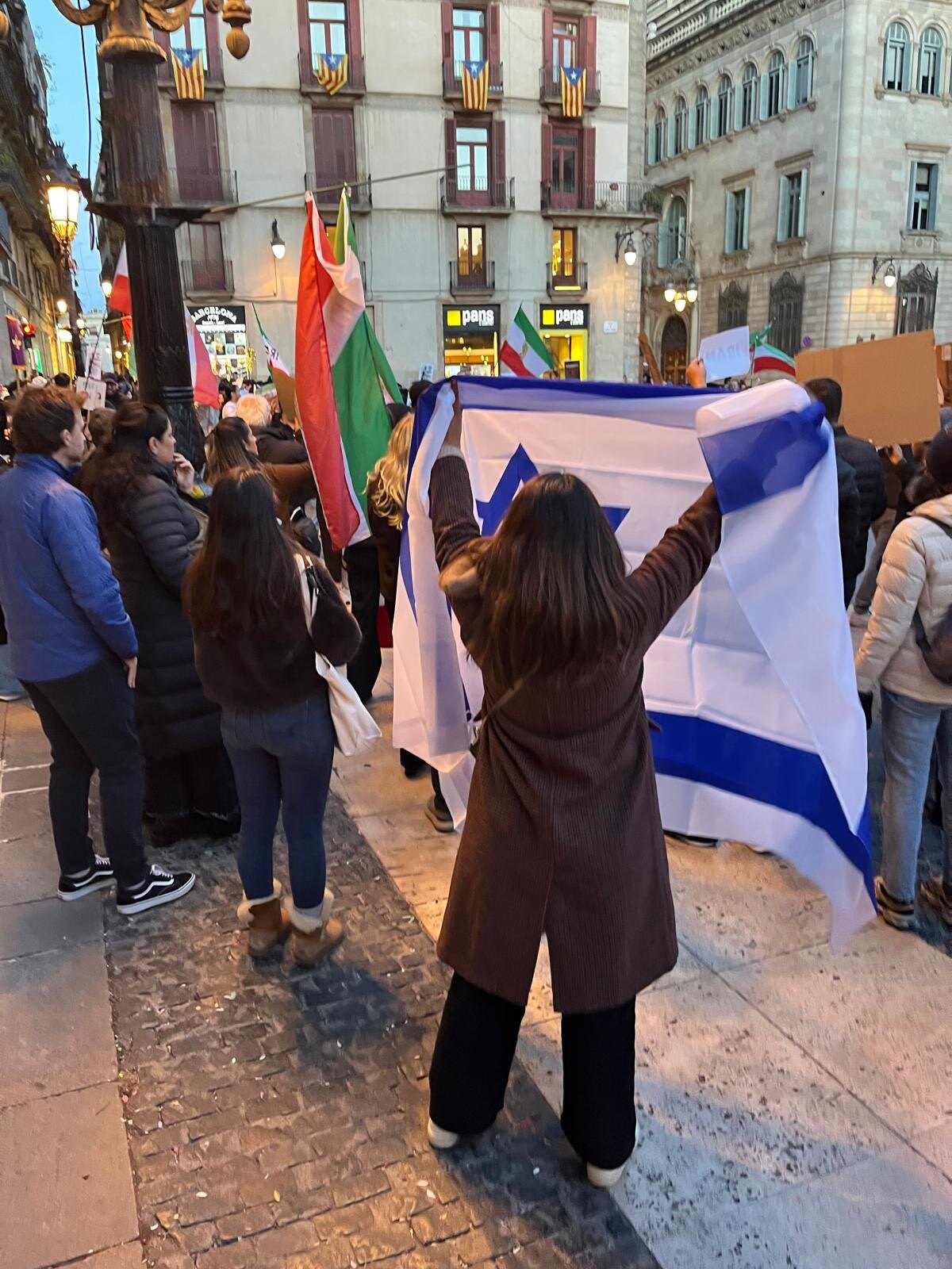 Al atardecer en la plaça de Sant Jaume, una participante despliega de espaldas una bandera de Israel por encima de su cabeza durante una concentración.